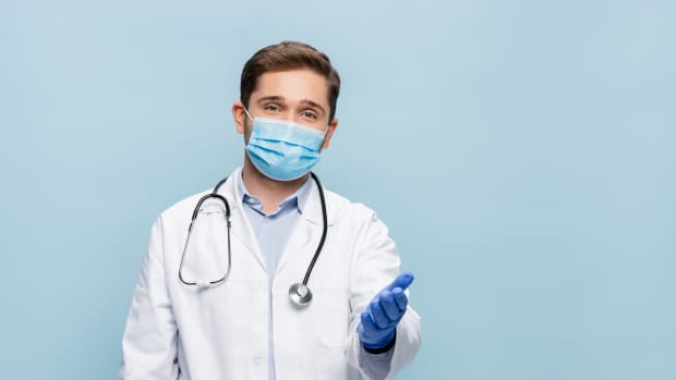 doctor in medical mask and white coat with stethoscope pointing with hand isolated on blue