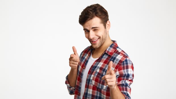 Close-up photo of playful shaven young man in checkered shirt pointing with two fingers, looking at camera, isolated on white background