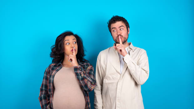 young couple expecting a baby standing against blue background silence gesture keeps index finger to lips makes hush sign. Asks not to share secret